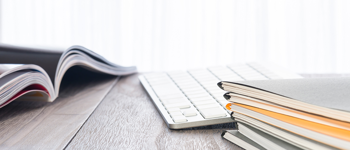 eine aufgeschlagene Broschüre und ein Stapel geschlossener Broschüren neben einer Tastatur auf einem Schreibtisch
