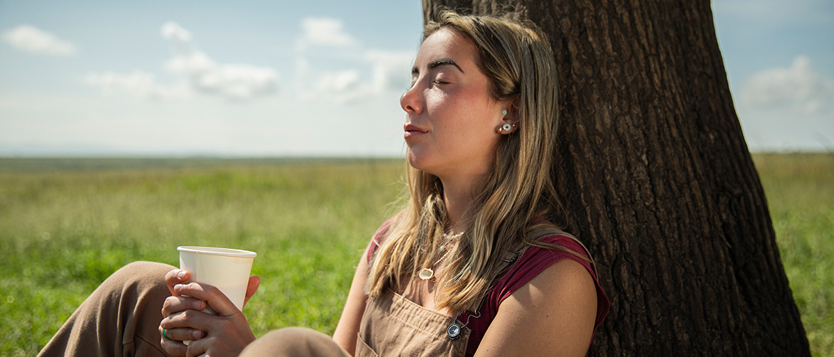 Frau sitzt an einem einsamen Baum mit einem Becher in der Hand.